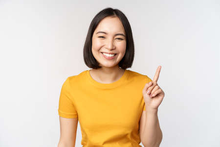 Beautiful young asian woman pointing finger up, smiling and looking amused at camera, showing advertisement, announcement on top, white backgroundの写真素材