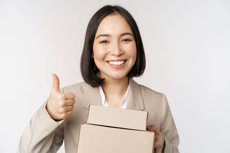 Smiling asian businesswoman, showing thumbs up and boxes with delivery goods, prepare order for client, standing over white backgroundの写真素材