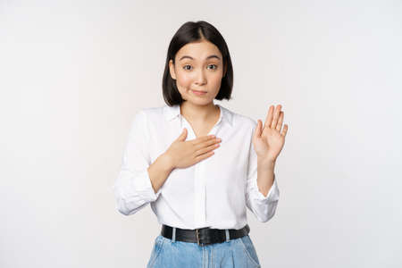 Image of cute young female office worker, asian girl student raising hand up and put palm on chest, name herself, introduce, making promise, standing over white backgroundの写真素材