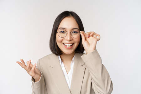 Enthusiastic asian saleswoman in glasses, smiling and laughing, looking amazed at camera, standing in beige suit over white backgroundの写真素材