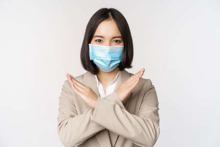 workplace concept. Image of asian businesswoman, female in medical face mask shows stop, cross prohibit gesture, stands over white backgroundの写真素材