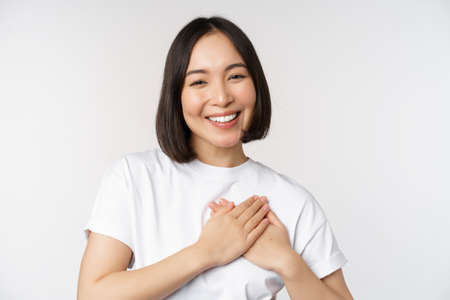 Close up of smiling korean woman holding hands on heart, care and love concept, feel affection, tenderness or heartwarming feeling, standing over white backgroundの写真素材
