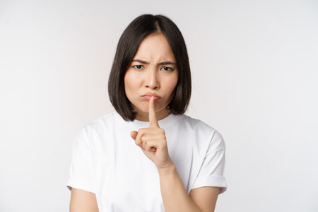 Angry asian girl shushing, keep quiet, taboo silence gesture, press finger to lips and frowning, scolding for being too loud, standing over white backgroundの写真素材