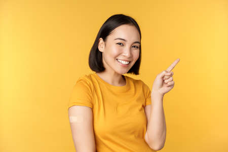 Vaccination and covid-19 pandemic concept. Smiling korean woman with strap on shoulder after coronavirus vaccine shot, pointing at banner with vaccinating campaignの写真素材