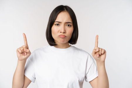 Sad and disappointed young asian woman pointing fingers up, showing advertisement with upset face...