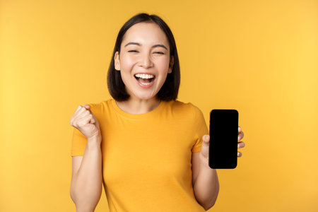 Excited asian woman, showing smartphone app and triumphing, celebrating on mobile phone, standing...
