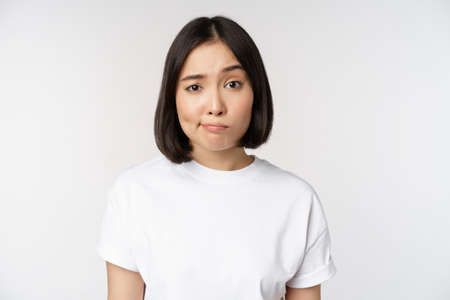 Image of young asian brunette woman in white t-shirt, smirk and look with doubt, hesitating, standing against white backgroundの写真素材