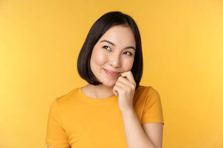 Close up portrait of cute asian girl smiling, thinking, looking up thoughtful, standing in tshirt over yellow backgroundの写真素材
