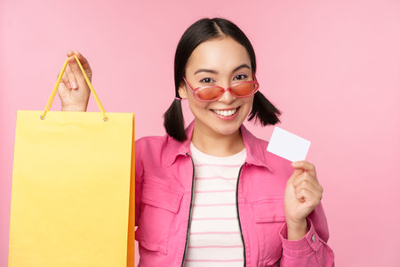 Happy young asian woman shows credit card and shopping bag, store sale announcement, buying smth in...