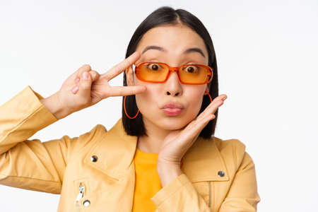 Close up portrait of young asian woman, stylish girl in sunglasses, showing peace, v-sign and smiling, standing over white studio backgroundの写真素材