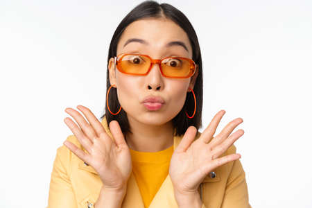 Stylish young asian woman in trendy sunglasses, pucker lips silly, looking surprised, impressed reaction, standing over white backgroundの写真素材