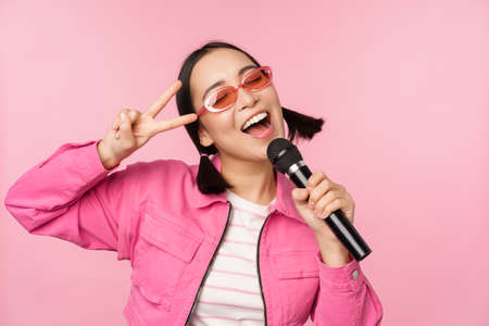 Happy beautiful asian girl singing with mic, using microphone, enjoying karaoke, posing against pink studio backgroundの写真素材