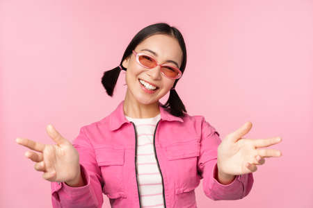 Happy stylish korean girl in sunglasses, reaching hands, receiving, smiling friendly and welcome you, standing over pink backgroundの写真素材