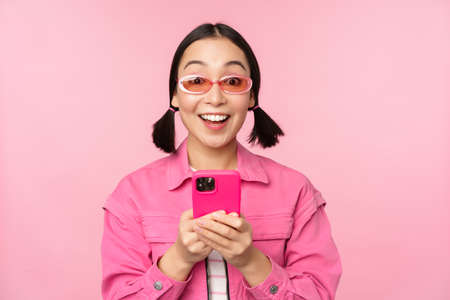Portrait of korean girl in stylish sunglasses holding mobile phone, using smartphone app, standing over pink backgroundの写真素材