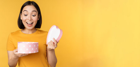 Portrait of excited asian woman, open gift box with surprised happy face, standing over yellow...