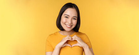 Close up portrait of smiling korean woman, showing romantic heart sign and looking happy, standing over yellow backgroundの写真素材
