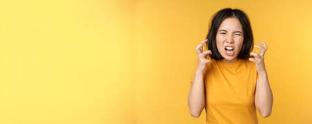 Image of angry asian woman, shouting and cursing, looking outraged, furious face expression, standing over yellow backgroundの写真素材