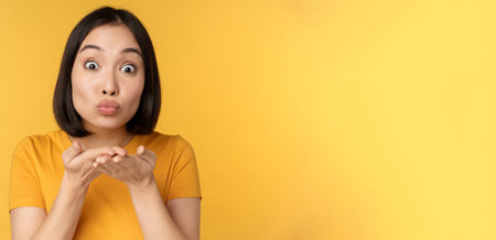 Cute asian girl sending air kiss, blowing mwah with puckered lips, standing over yellow background