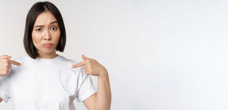 Young asian woman pointing at herself with disbelief, being chosen, surprised by her candidature,...