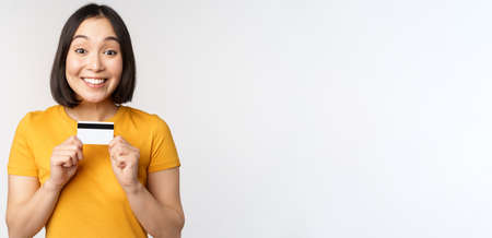 Portrait of beautiful korean girl holding credit card, recommending bank service, standing in yellow tshirt over white backgroundの写真素材