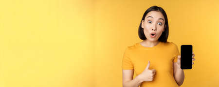 Excited asian woman showing mobile phone screen, thumbs up, like smth good, recommending smartphone application, standing over yellow backgroundの写真素材
