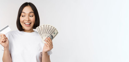 Portrait of asian woman smiling, holding credit card and money cash, dollars, standing in tshirt...