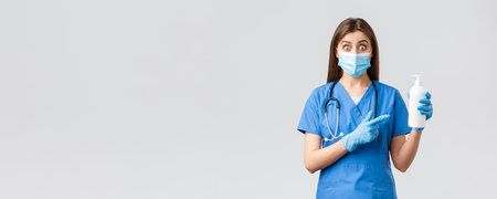 Covid-19, preventing virus, health, healthcare workers and quarantine concept. Surprised female nurse, cute doctor in blue scrubs, medical mask and gloves pointing finger at hand sanitizer or soapの写真素材