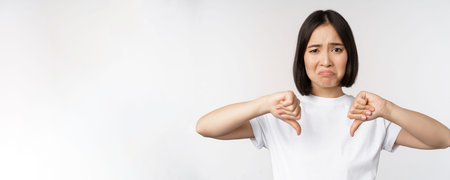 Image of asian woman showing thumbs down, dislike smth, looking disappointed, standing over white...