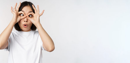 Funny young asian woman, korean girl making eyes glasses gesture, looking happy at camera, standing...