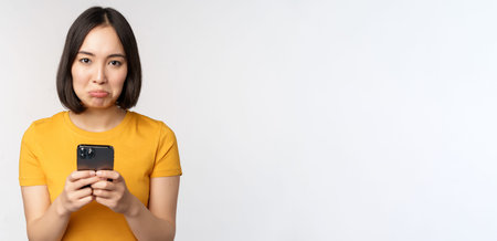 Sad asian woman holding smartphone, looking upset with regret, standing in yellow tshirt against...