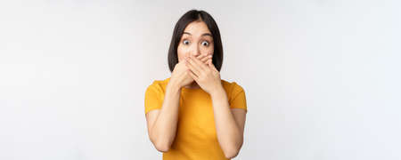 Shocked asian woman cover mouth with hands, looking startled with speechless face expression, standing in yellow tshirt against white backgroundの写真素材