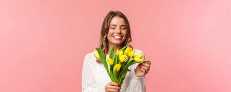 Spring, happiness and celebration concept. Close-up portrait of lovely romantic smiling girl touching petal of yellow tulip, holding flowers, receive bouquet on date, standing pink backgroundの写真素材