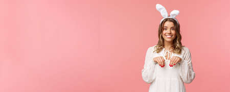Holidays, spring and party concept. Portrait of cute and tender, lovely blond girl imitating bunny, holding hands like paws and wearing rabbit ears, smiling camera, pink backgroundの写真素材