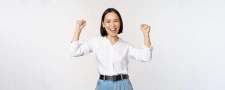 Enthusiastic asian woman rejoicing, say yes, looking happy and celebrating victory, champion dance, fist pump gesture, standing over white backgroundの写真素材