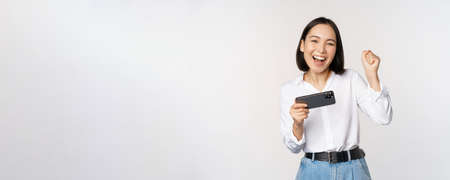 Happy asian woman do winner dance, triumphing, winning on mobile phone video game, holding smartphone horizontal position and celebrating, white backgroundの写真素材
