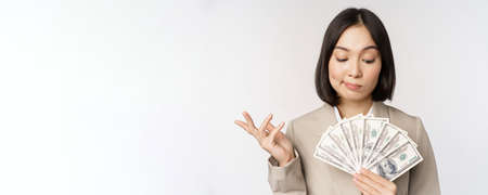 Image of asian corporate woman, happy businesswoman showing money, cash dollars and thinking, standing in suit over white backgroundの写真素材