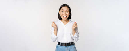 Enthusiastic asian woman rejoicing, say yes, looking happy and celebrating victory, champion dance, fist pump gesture, standing over white backgroundの写真素材