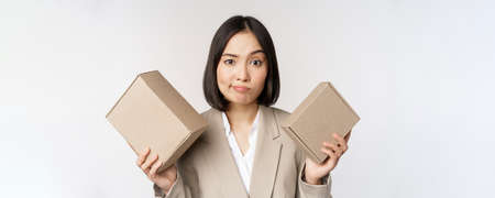 Puzzled asian saleswoman holding boxes, looking confused, standing in business suit over white backgroundの写真素材