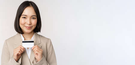 Smiling office clerk, asian corporate woman showing credit card, standing over white background in beige suitの写真素材