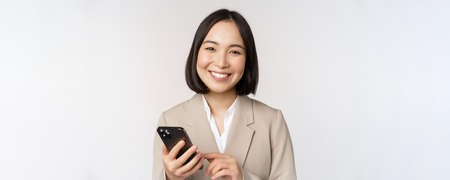 Close up portrait of korean woman, corporate lady in suit, using mobile phone and smiling, holding smartphone, standing over white backgroundの写真素材