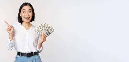 Smiling young modern asian woman, pointing at banner advertisement, holding cash money dollars, standing over white backgroundの写真素材