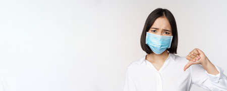 Portrait of asian woman in medical face mask showing thumbs down with disappointed, tired face expression, standing over white backgroundの写真素材