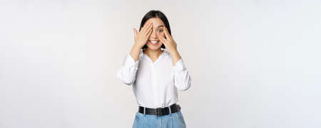 Image of young asian woman close her eyes and smiles, waits for surprise, anticipates, peeking through fingers, stands over white backgroundの写真素材
