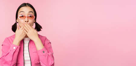 Portrait of shocked, stylish asian girl in sunglasses, closes mouth, looks with surprised face expression, stands over pink backgroundの写真素材