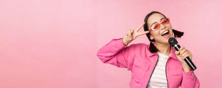 Happy beautiful asian girl singing with mic, using microphone, enjoying karaoke, posing against pink studio backgroundの写真素材