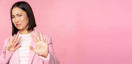 Get away. Disgusted asian businesswoman rejecting, shaking hands to refuse, grimacing from dislike, decline smth with aversion, posing in suit against pink backgroundの写真素材