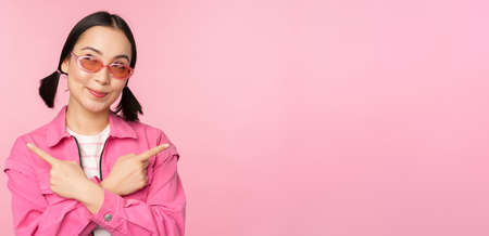 Choice. Stylish korean girl, asian female model points fingers sideways, shows two variants, product advertisement, demonstrating items, standing over pink backgroundの写真素材