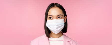 Close up portrait of japanese corporate woman in medical face mask from covid-19, looking left at logo, sale promo, standing over pink backgroundの写真素材