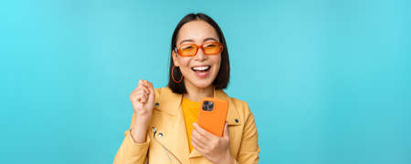 Happy stylish asian girl using smartphone and laughing, smiling at camera, standing over blue backgroundの写真素材