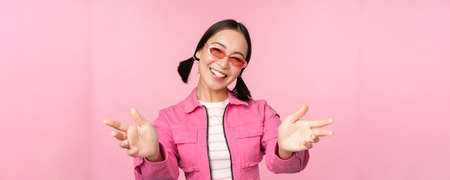 Happy stylish korean girl in sunglasses, reaching hands, receiving, smiling friendly and welcome you, standing over pink backgroundの写真素材
